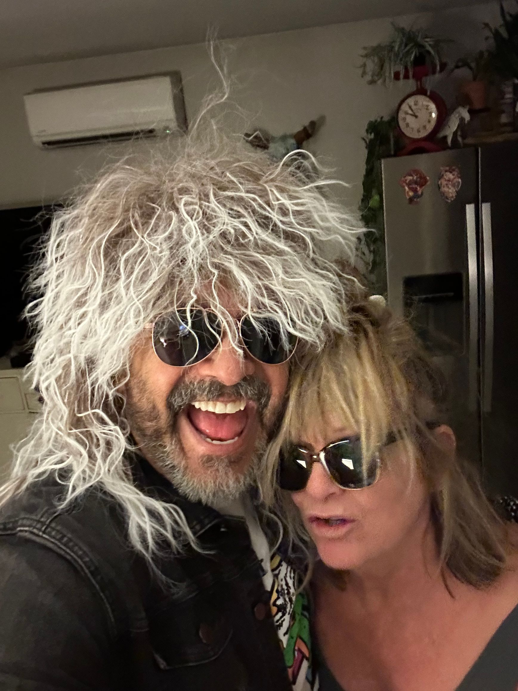 A close-up selfie of two people wearing sunglasses, with one wearing a long, grey, textured wig and smiling broadly.