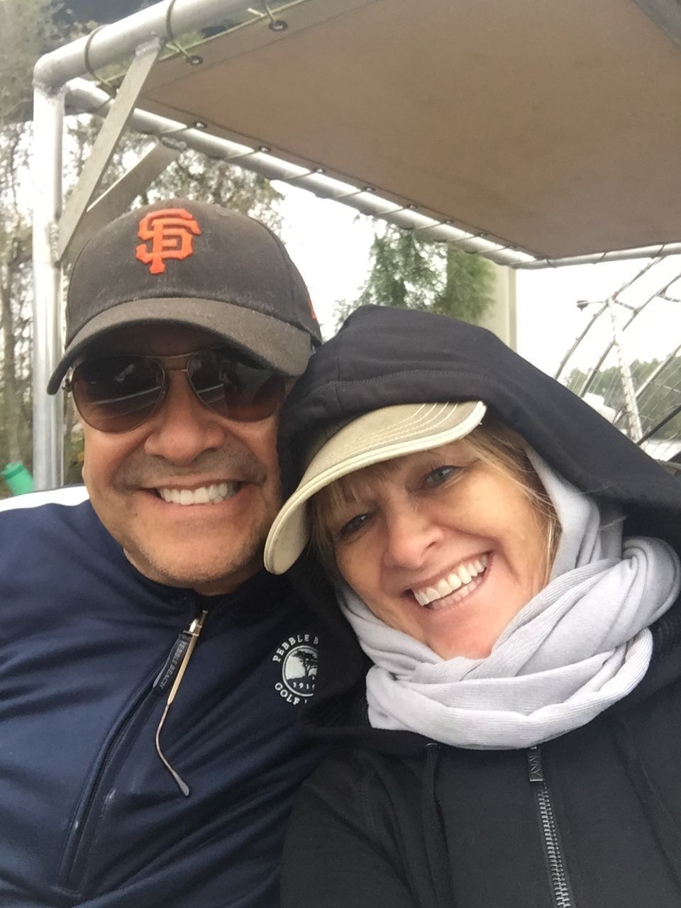 A man wearing a baseball cap and sunglasses and a woman in a hooded jacket and scarf smiling together outdoors.