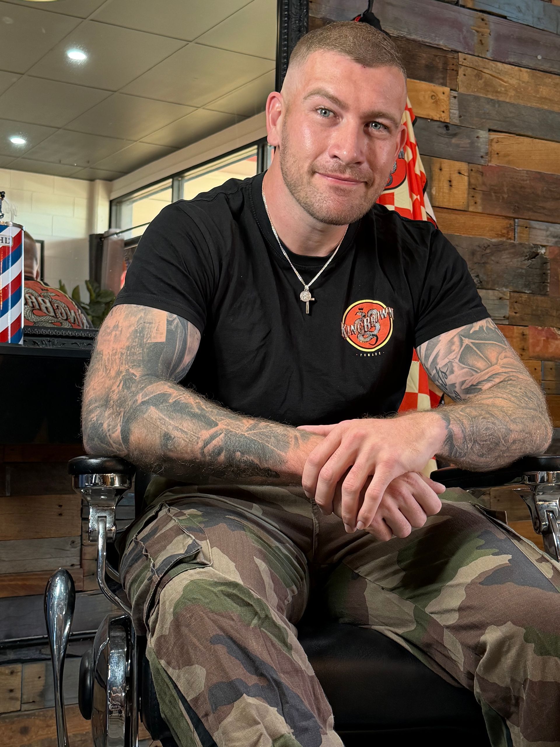 Tattooed man in a black shirt and camouflage pants sitting indoors, arms crossed and smiling lightly