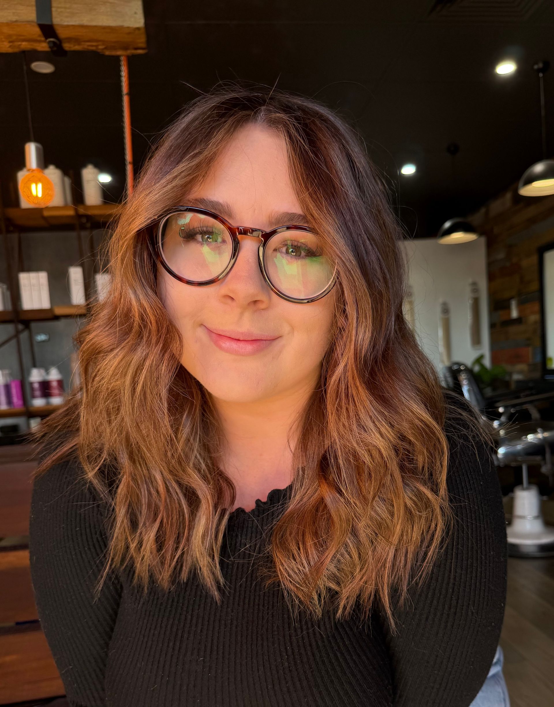 Person with wavy hair and round glasses, smiling in a warmly lit cafe.