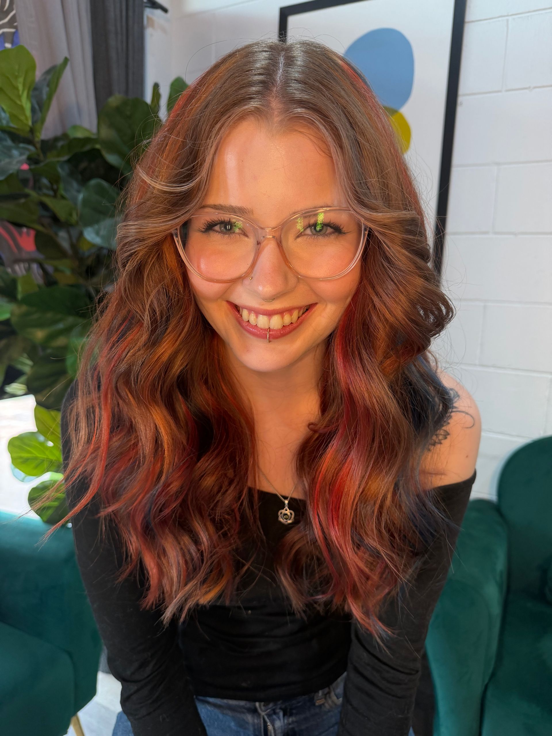 Smiling person with glasses and wavy reddish hair seated on a green couch indoors