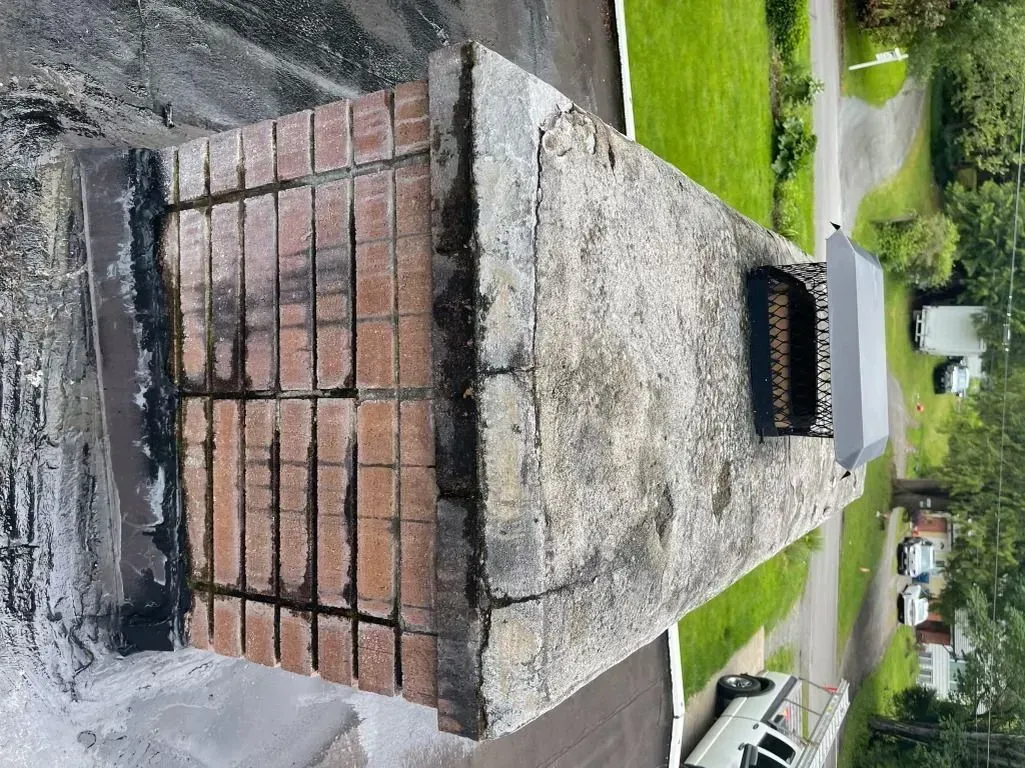 Chimney on a roof with brick sides, a concrete top, and a metal cap, with a background of green grass and houses.