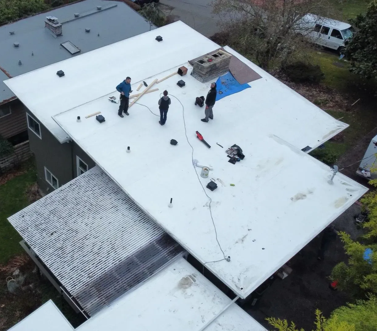 Aerial view: Workers on a white flat roof, installing something. Houses and trees surround.