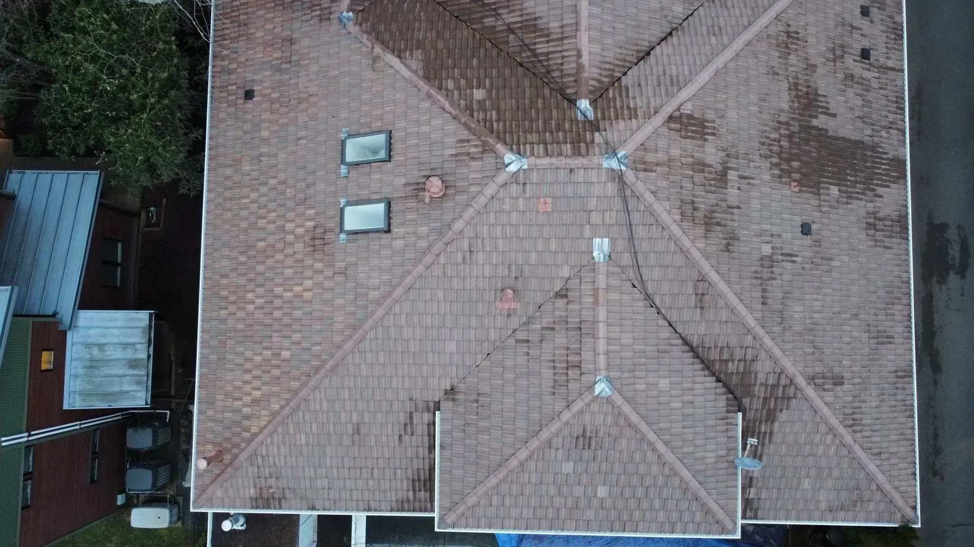 Overhead view of a brown shingled house roof with two skylights, vents, and surrounding cityscape.