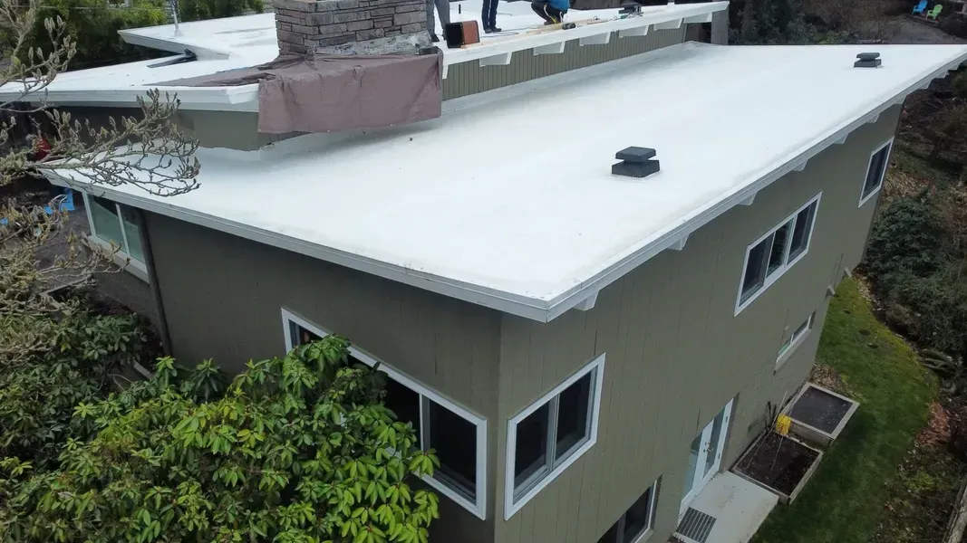 A light-colored flat roof on a two-story house, with workers on the roof and green siding.