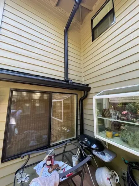 Exterior corner of a beige building with black gutters. A window and patio furniture are visible.