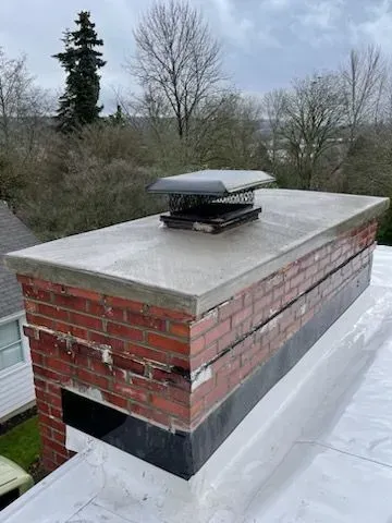 Brick chimney on a white roof, with a metal cap.  Cracked bricks visible. Overcast sky, trees in background.