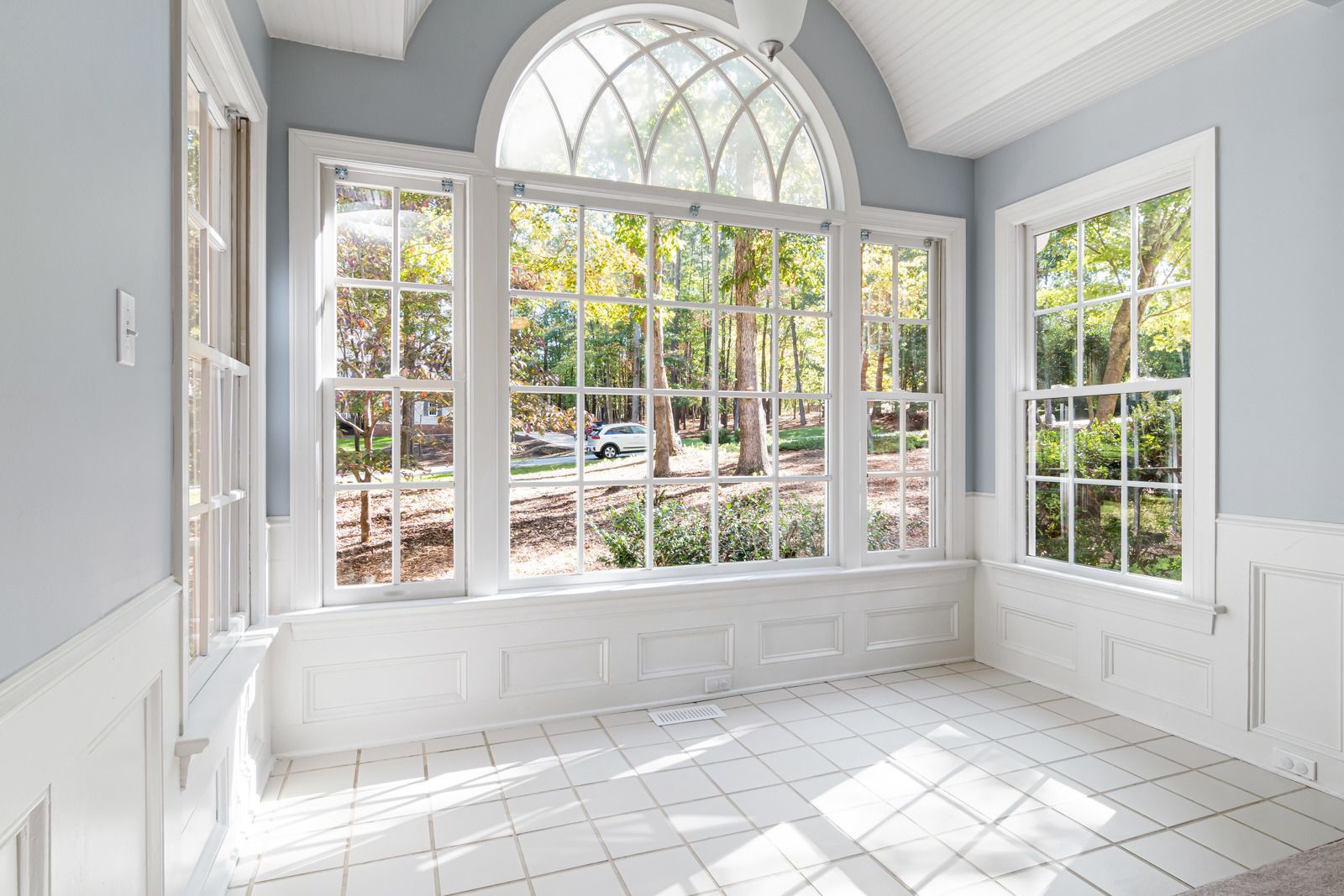 Sunroom with large white windows, light blue walls, and white tile floor overlooking trees.