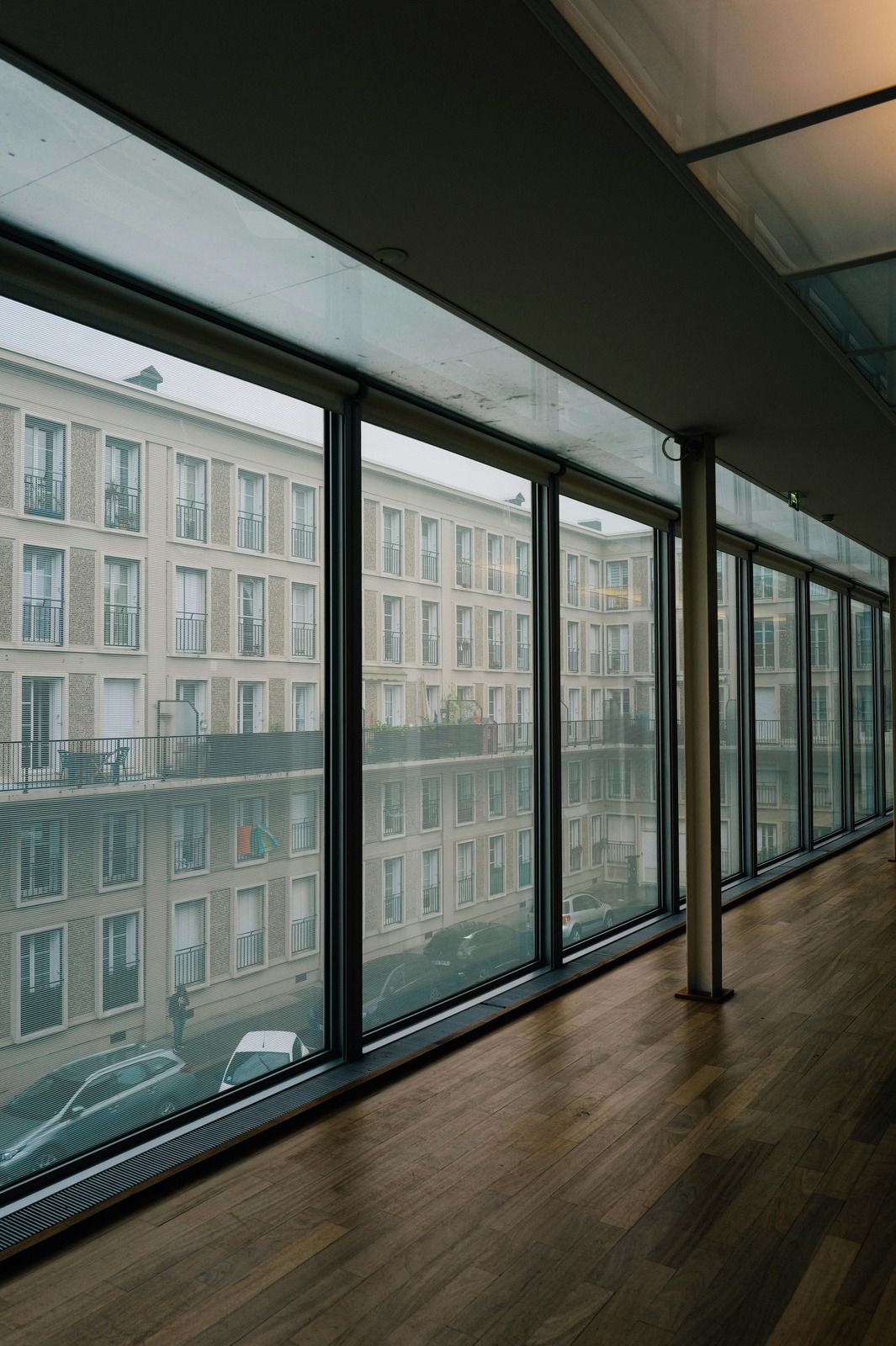 A long interior view with large windows overlooking an apartment building and a wooden floor.