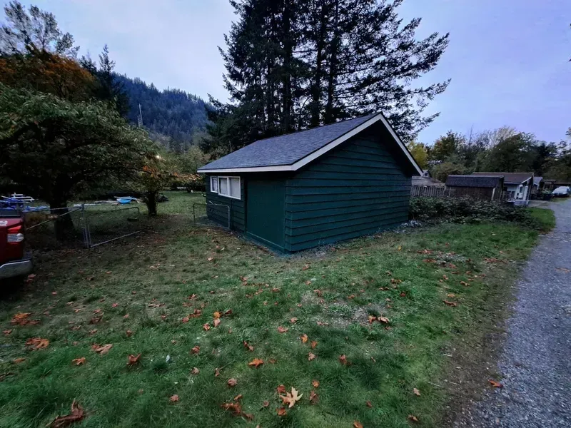 A green, rectangular shed with a dark roof sits on a grassy lawn. A car is on the left.