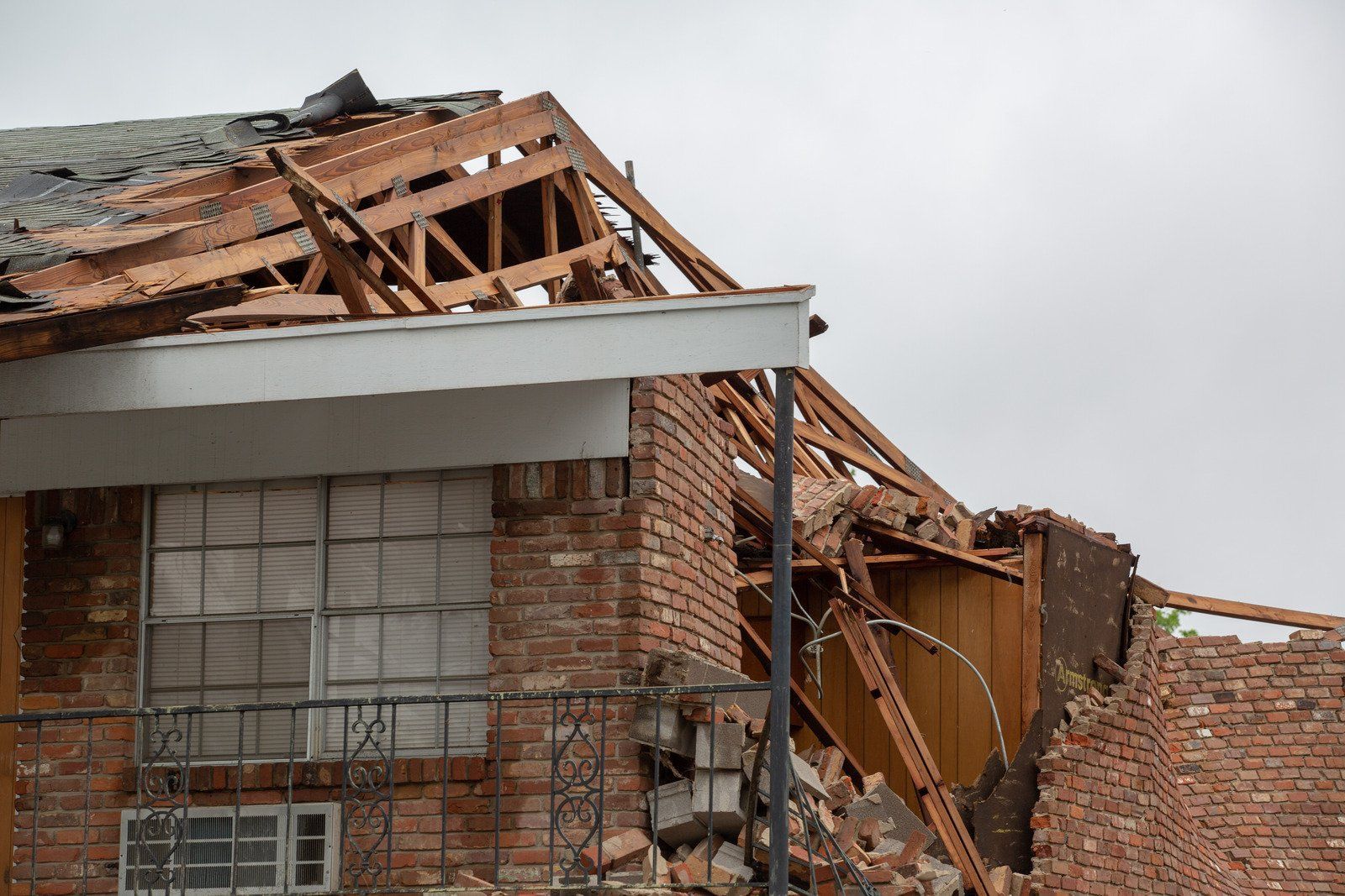 A brick building with a destroyed roof, debris scattered, under a cloudy sky.