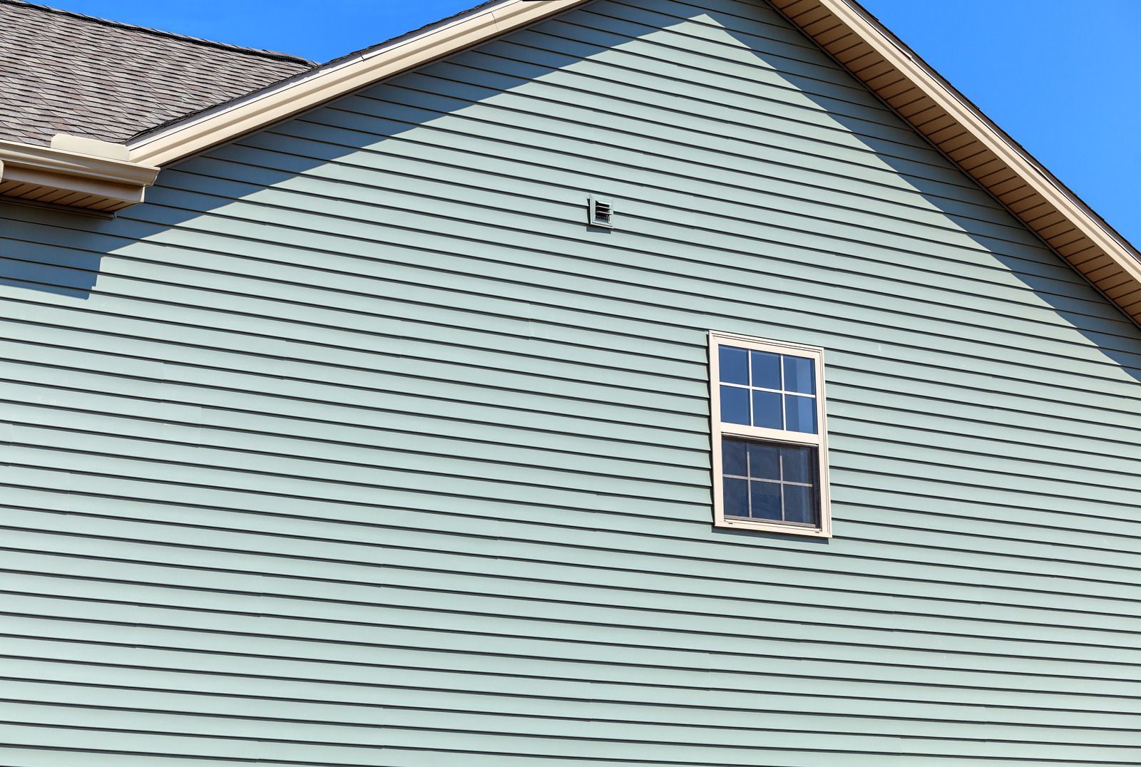 Blue siding on a house with a window and a brown roof against a blue sky.
