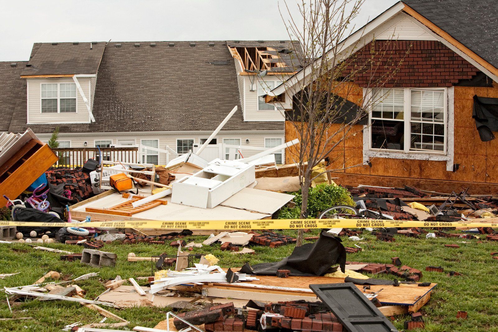 House damaged by a storm, debris scattered across the yard, caution tape visible.