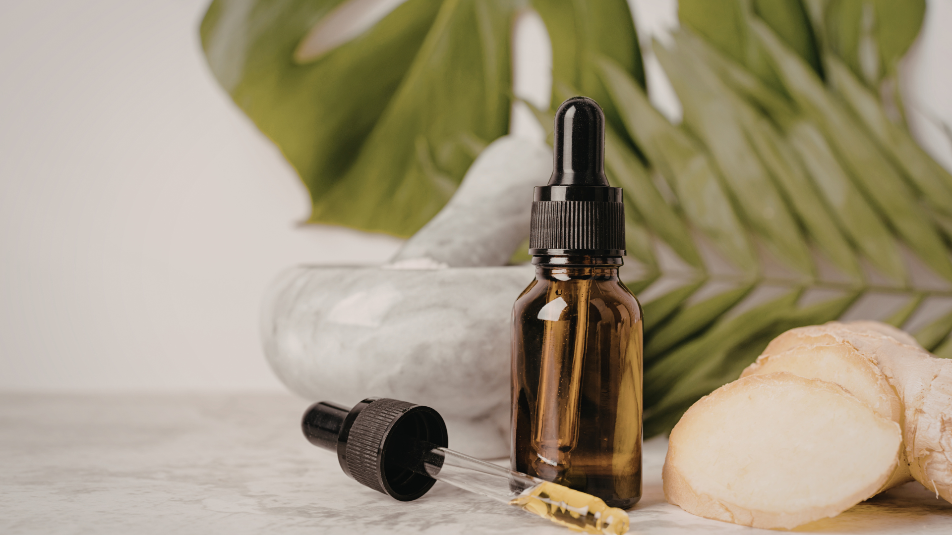 Amber essential oil bottle with dropper, mortar, sliced ginger, and Monstera leaf on a white surface.