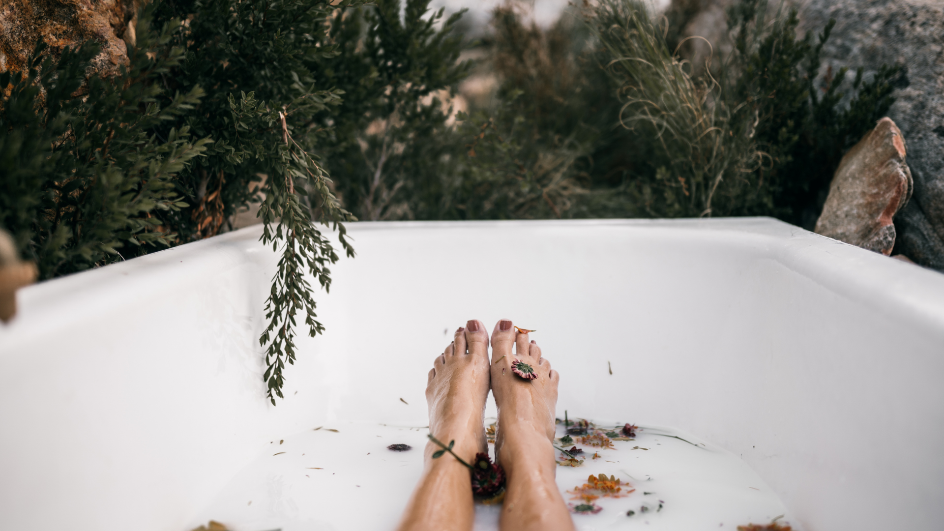 Feet in a white bathtub with milky water and leaves, surrounded by plants and rocks.