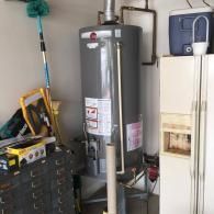 A gray Rheem water heater in a cluttered garage, positioned next to a tall white cabinet and various tools.