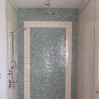 A walk-in shower with light blue mosaic tiles, framed by white tiles, featuring a chrome shower head and fixtures.