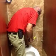 A person in a red shirt and tan pants repairs a wall-mounted fixture above a toilet in a stall with tiled walls.