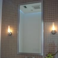 A tall, rectangular mirror with a silver frame is mounted on a wall with brown, square mosaic tiles and two wall sconces.