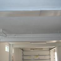 An interior view of a garage with white painted walls, a concrete ceiling with exposed pipes, and a metal roll-up door.