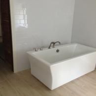 A modern white rectangular freestanding bathtub with chrome fixtures sits against a white tiled wall on a wood floor.