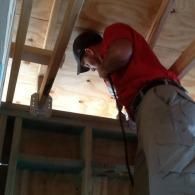 A person in a red shirt and baseball cap looking upward at wooden ceiling joists and plywood in a construction setting.