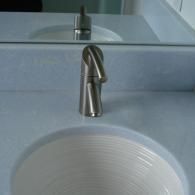 A modern brushed metal faucet centered above a white, circular bathroom sink set in a light grey countertop.
