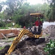 A person in a red shirt operates a yellow mini excavator to dig a trench in a wooded residential yard.