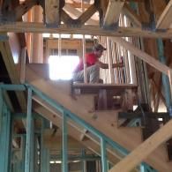 A worker in a red shirt installs railings on wooden stairs inside a house under construction.