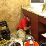 A worker in a red shirt and tan pants kneels on the floor, fixing plumbing under a bathroom vanity.