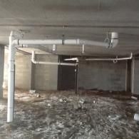 A network of white PVC plumbing pipes suspended from a concrete ceiling in an unfinished building interior.