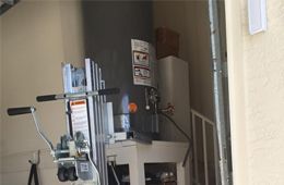 A hand truck stands in front of a dark gray water heater mounted on a white platform inside a garage or utility area.