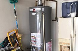 A grey Rheem water heater standing in a utility area with household tools and a cooler nearby.
