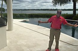 A smiling person in a red polo shirt and khakis stands with arms outstretched on a patio by a pool overlooking a lake.
