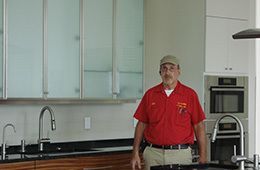 A person in a red work shirt and cap stands in a modern kitchen with frosted cabinets and dark countertops.