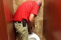A person wearing a red shirt and tan pants is leaning over to repair a toilet in a tiled bathroom stall.