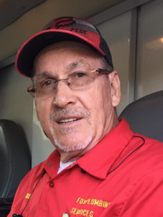 A person wears a red collared shirt with yellow text and a red and black baseball cap while smiling.
