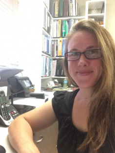 A person with long hair and glasses smiles while sitting at a desk in an office with bookshelves in the background.