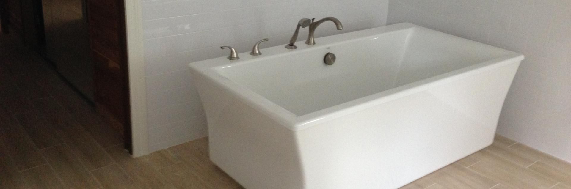 A white, rectangular freestanding bathtub with a silver faucet and handles sits on a light wood floor against white walls.