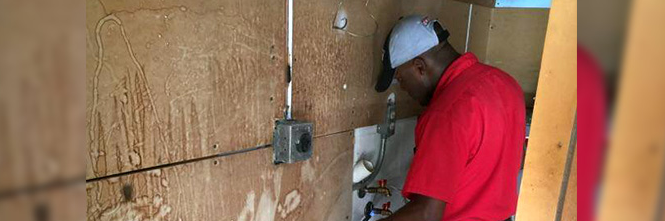 A person in a red shirt and gray cap working on plumbing behind a wooden wall with an electrical box.