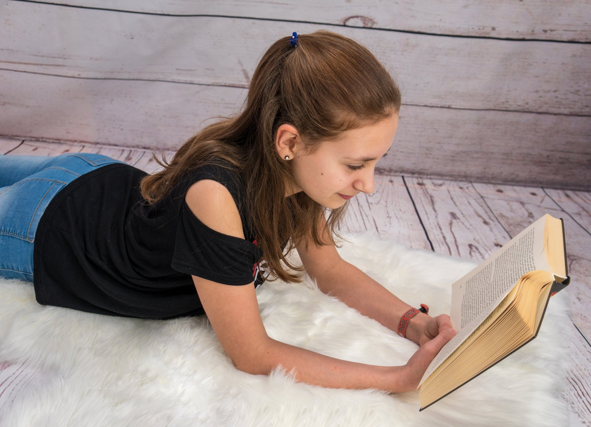Kind Teeanager Mädchen Buch Fell Teppich braun Armband lesen liegen Holz Hintergrund grau
