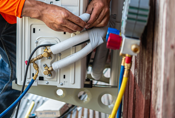 Technician connecting white insulated pipes and colored hoses to an outdoor air conditioner unit