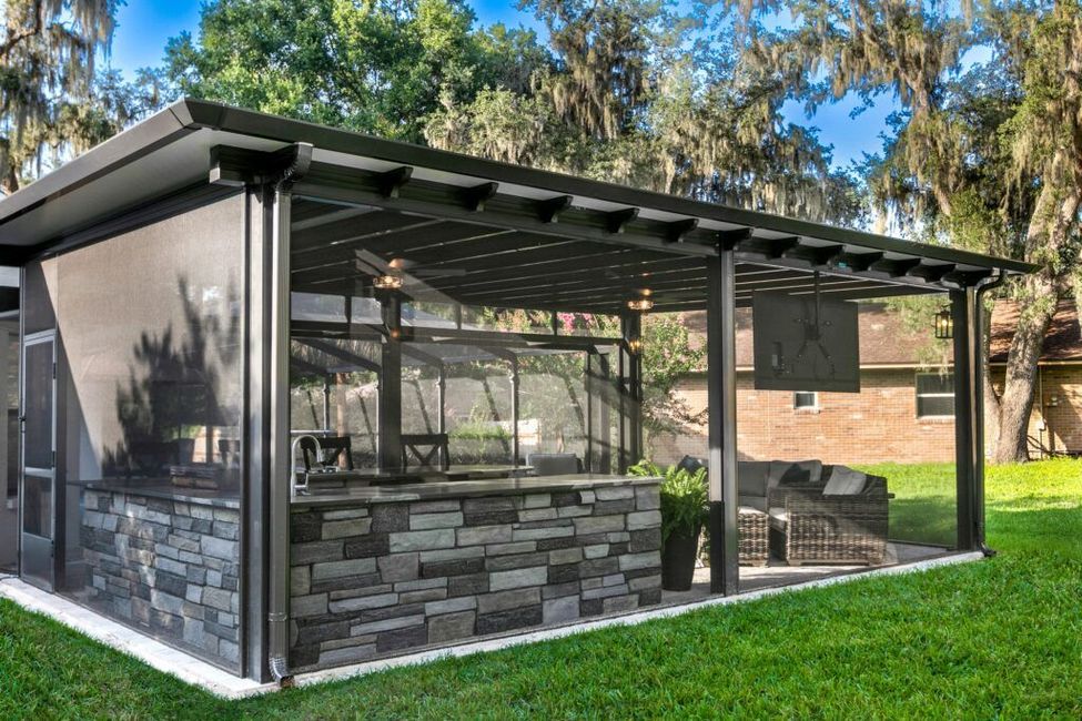 Outdoor screened-in kitchen with stone bar, seating area, and TV under a dark-colored roof, surrounded by trees.