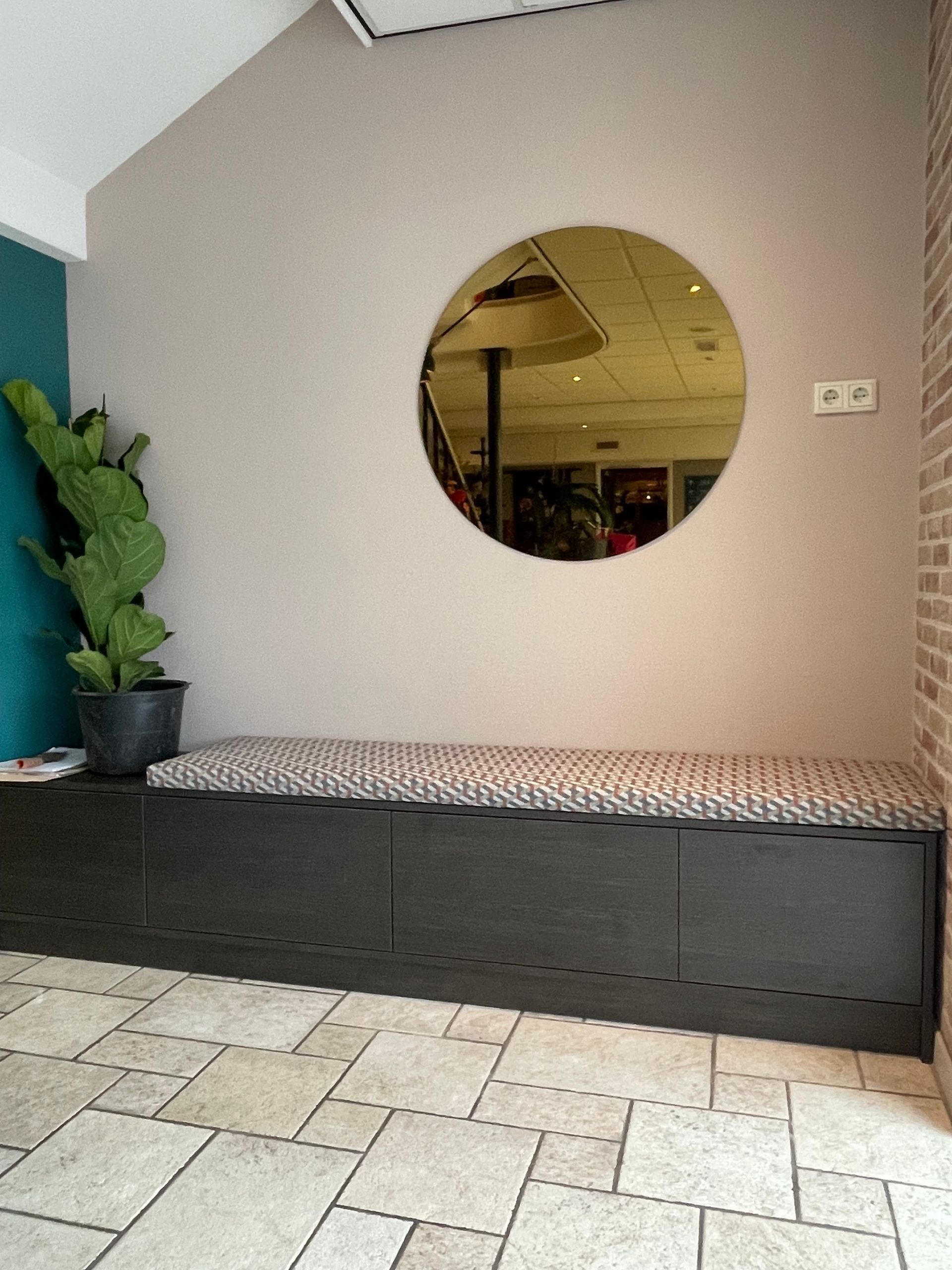 Bankje met kussen met patroon onder een ronde spiegel, naast een cactus en een turquoise muur.