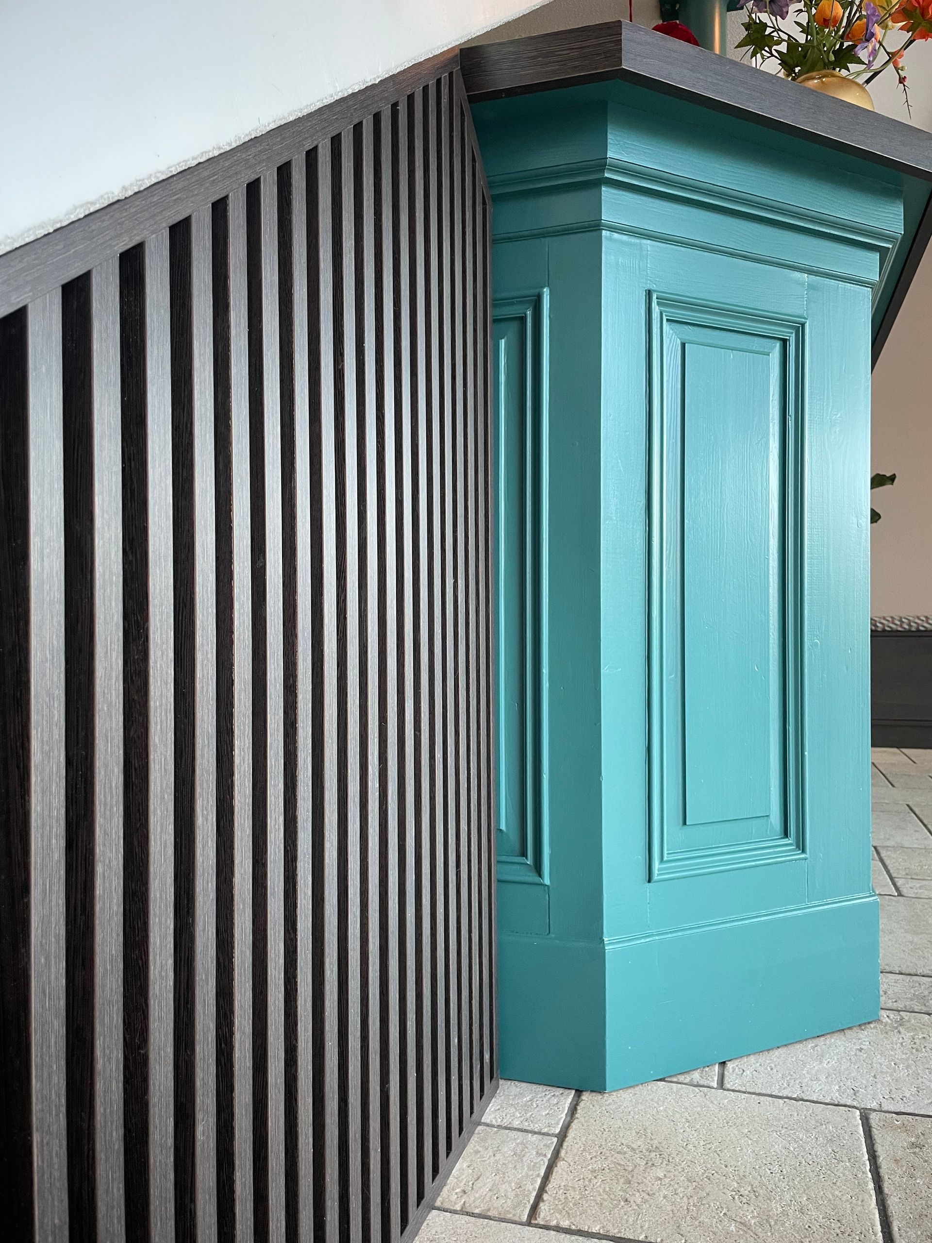 Een turkooizen geschilderde kolom naast een zwarte lattenwand, hoekdetail. Grijze tegelvloer.