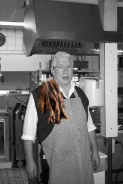 Een man met een schort met bontrand staat in een professionele keuken met roestvrijstalen apparatuur.