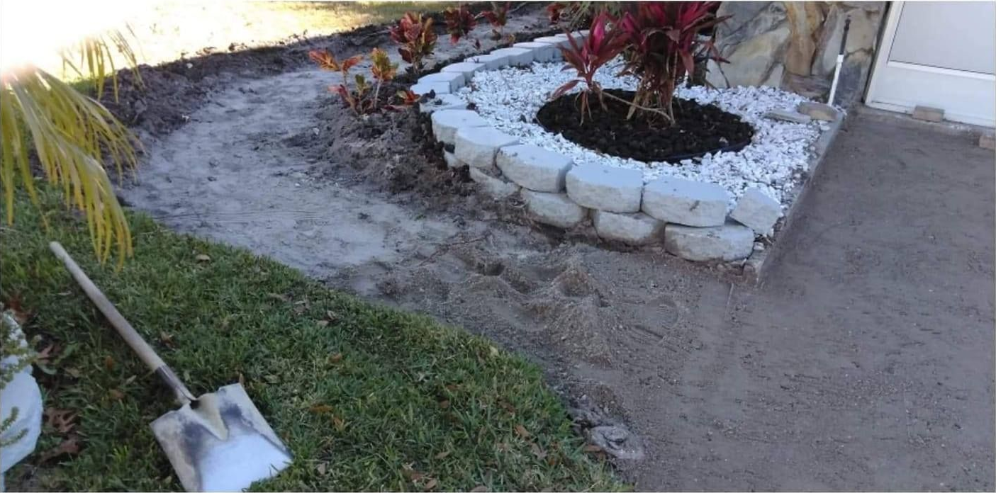 A garden bed with plants, white stones, and a dirt path. A shovel lies on the green grass.