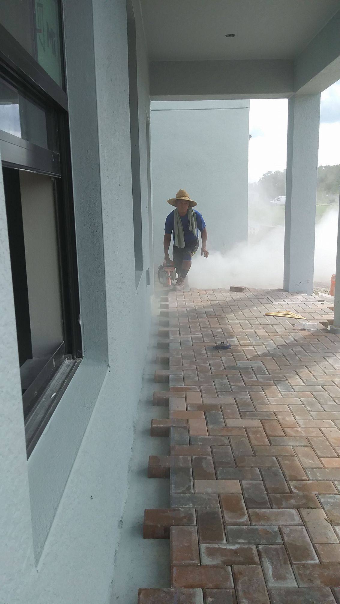 Person in a straw hat pushing a wheelbarrow on a brick pathway, creating a dust cloud near a building.