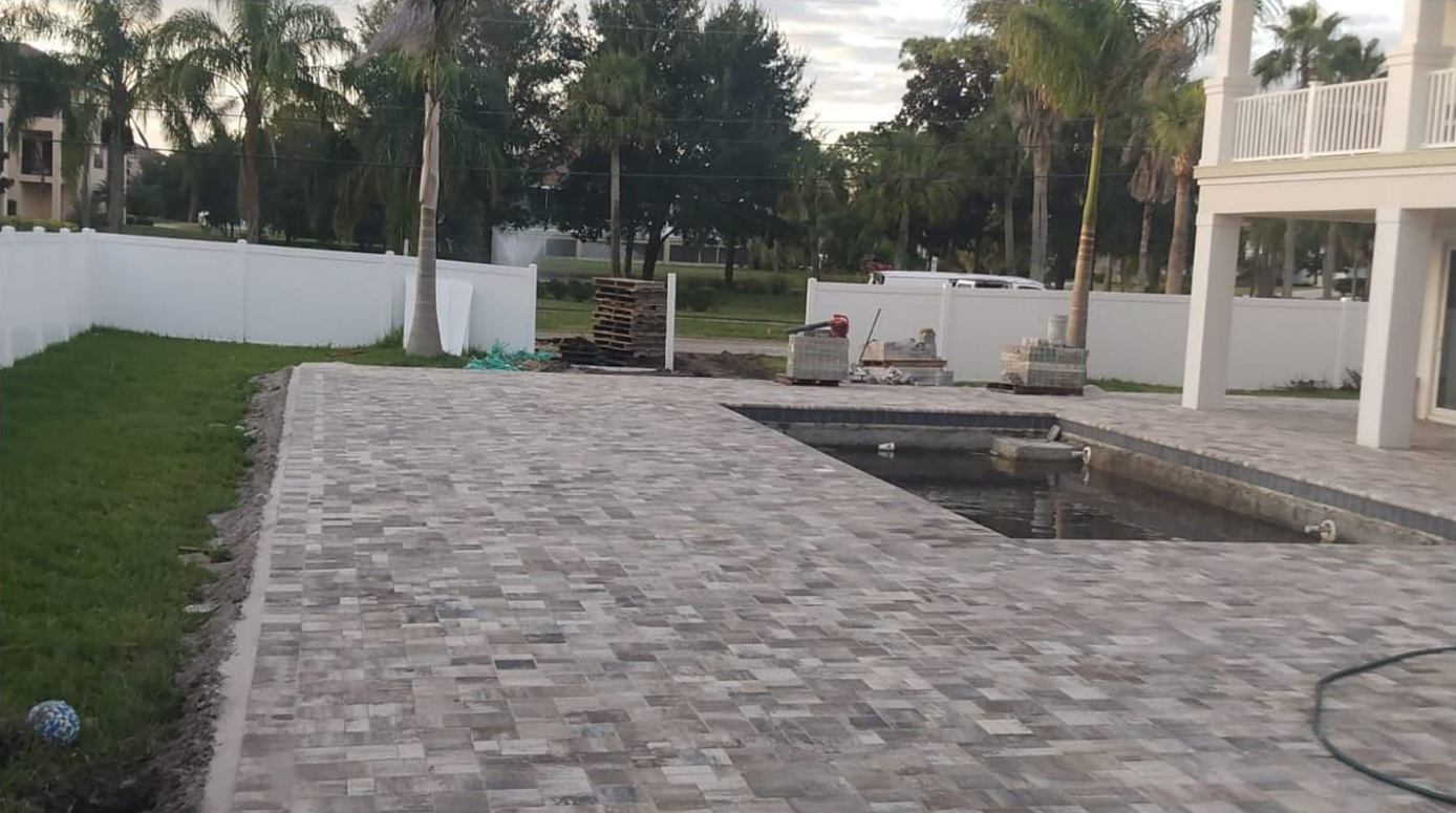 Patio with brick pavers, pool, and white fence in a backyard setting.