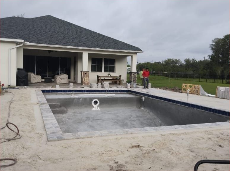 Pool construction in progress near a house with a man standing nearby. Pool is empty and gray, house is light-colored.